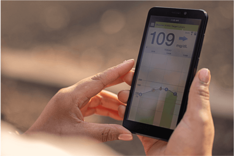 Hände halten im Freien ein Smartphone, das den Glukoseverlauf über die Zeit anzeigt und den Glukosewert „109 mg/dL“.