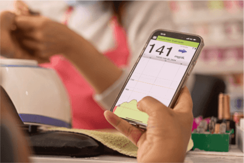 Hand hält ein Smartphone, das mit der Eversense-App den Glukosewert „141 mg/dL“ anzeigt.